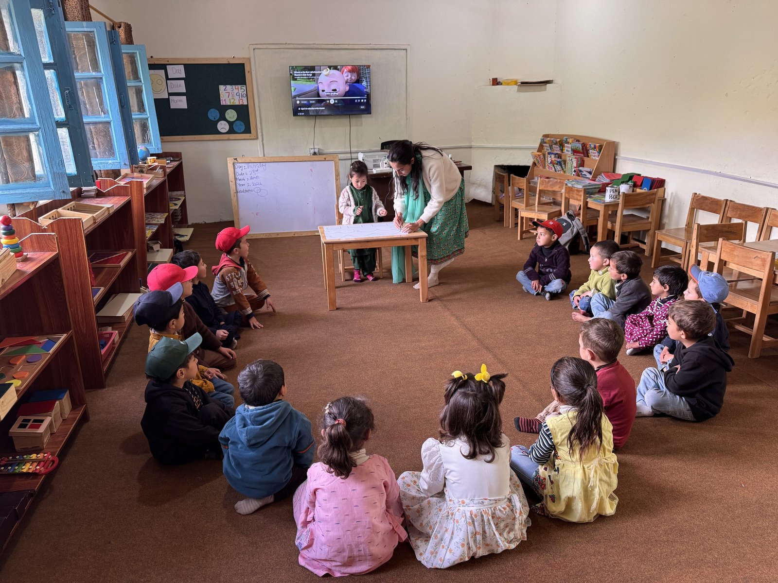 A teacher guides a student through early writing practice while classmates watch.