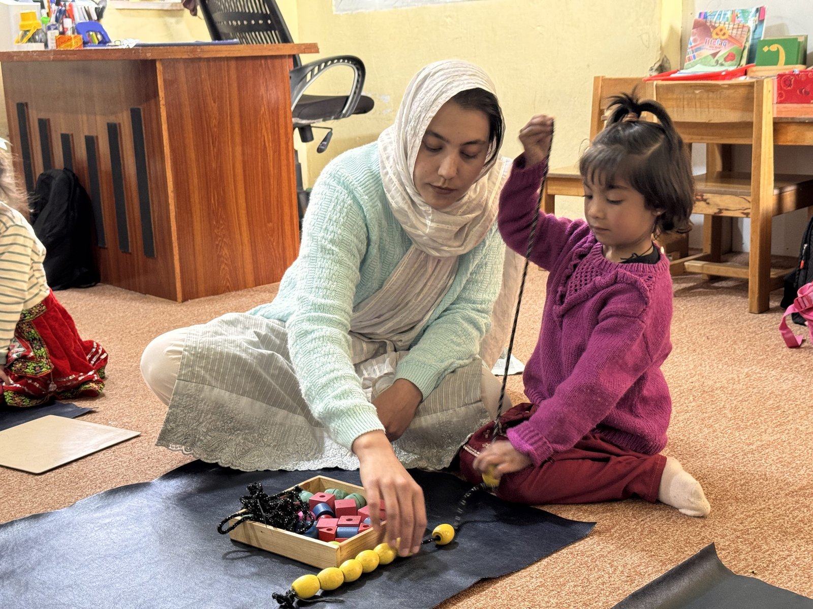 A teacher works one-on-one with a student on a threading and beading activity.